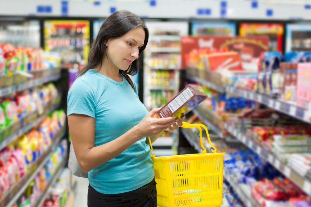 A woman in a supermarket aisle reading the back of a product label while holding a yellow shopping basket.