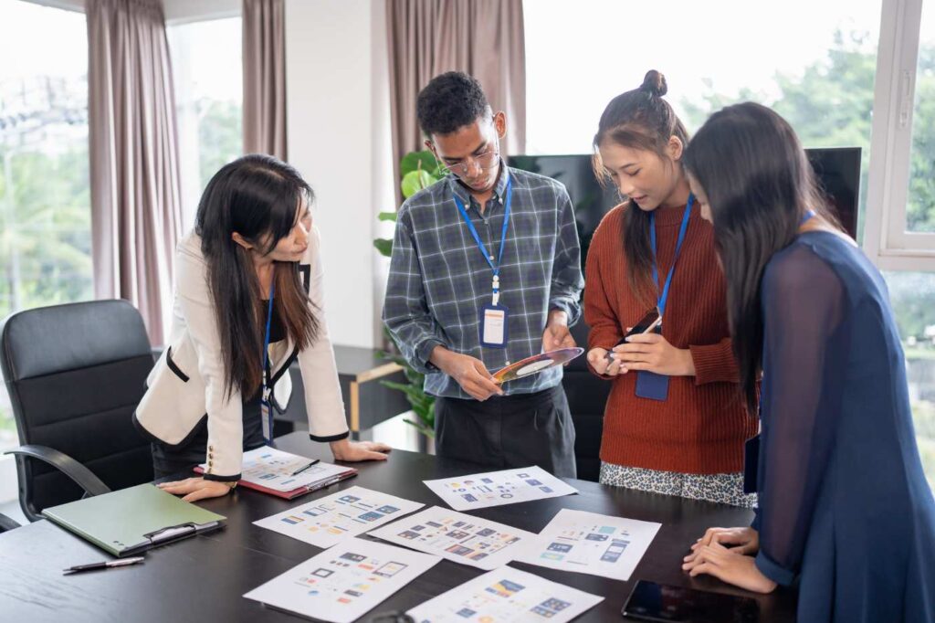 A diverse team standing around a table reviewing printed product layouts and packaging designs.