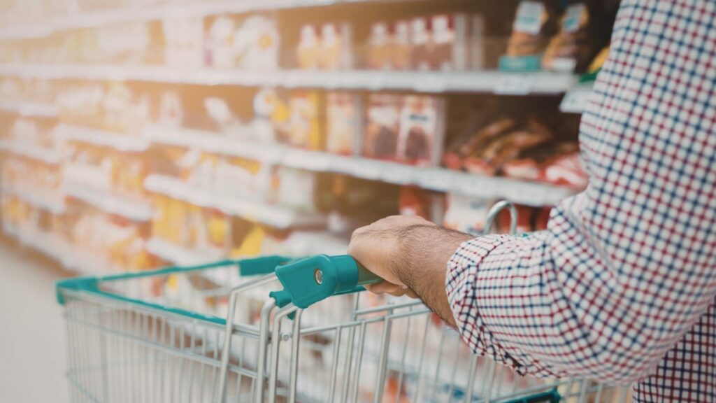 Person pushing a shopping trolley through a supermarket aisle, browsing packaged food.