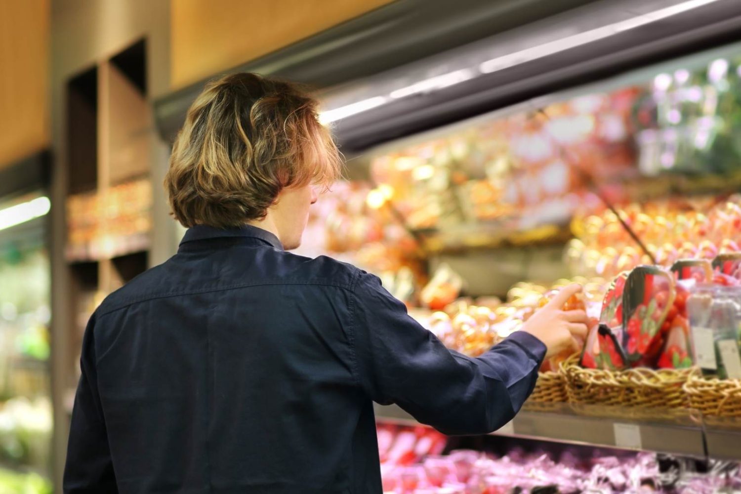 A shopper selecting items from a refrigerated supermarket shelf filled with fresh produce and prepared foods.