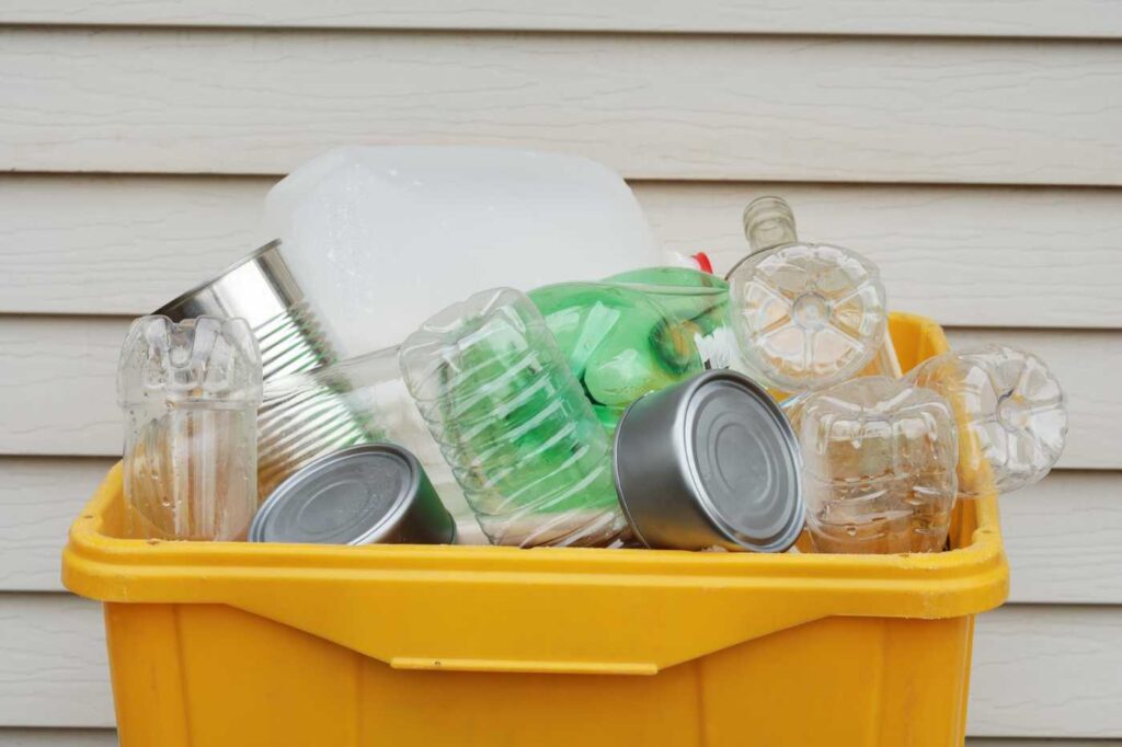 A yellow recycling bin filled with various plastic bottles, containers, and aluminum cans.